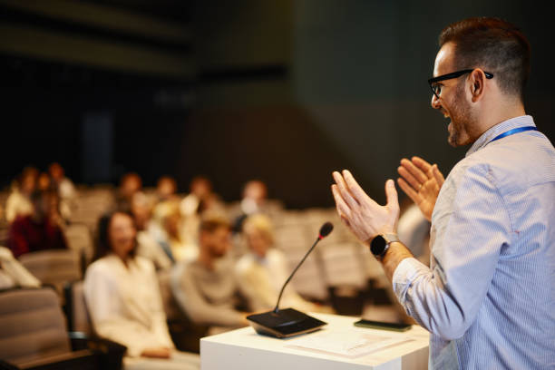 Young happy businessman giving a speech in front of large group of his colleagues at convention center. Copy space.